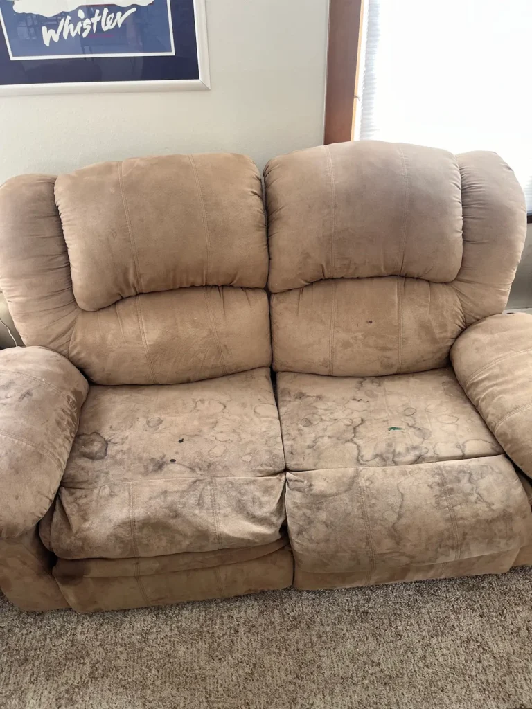 A visibly dirty brown suede loveseat sofa with lots of dark stains.